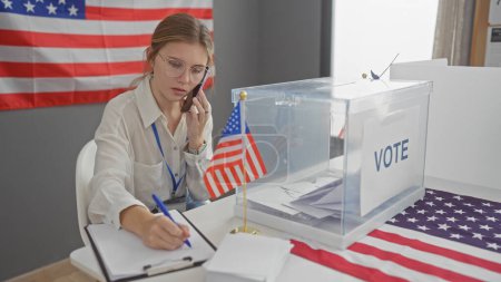 Photo for A young caucasian woman multitasks, writing and speaking on the phone at a us electoral voting center with flags - Royalty Free Image