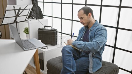 Ein glatzköpfiger hispanischer Mann mit Bart schreibt im Haus Notizen und reflektiert seine Arbeit am Laptop und Notenpult..