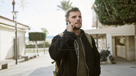 Ein junger hispanischer Mann mit Bart telefoniert auf einer Straße in der Stadt und beschreibt ein urbanes Ambiente im Freien.