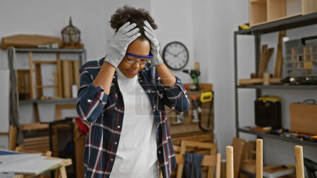 Stressed woman with curly hair wearing safety goggles in workshop