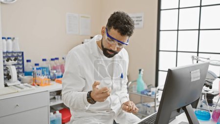 Foto de Un hombre barbudo examina una botella mientras trabaja en una computadora portátil en un entorno de laboratorio, simbolizando la investigación y la atención médica. - Imagen libre de derechos