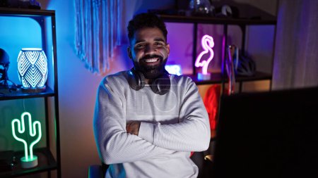 A young hispanic male smiles with crossed arms in a neon-lit gaming room at night, exuding a trendy and relaxed vibe.
