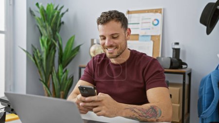 Hombre hispano sonriente con barba usando teléfono inteligente en un entorno de oficina moderno