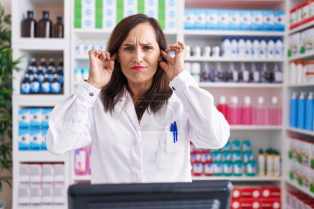 Photo for Middle age brunette woman working at pharmacy drugstore covering ears with fingers with annoyed expression for the noise of loud music. deaf concept. - Royalty Free Image