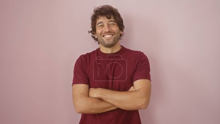 Handsome hispanic man with a beard smiling with arms crossed against a pink wall in a casual maroon shirt.
