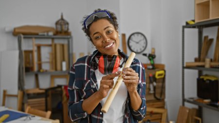 Téléchargez les photos : Une femme heureuse aux cheveux bouclés portant des lunettes de sécurité et des cache-oreilles tient des outils de travail du bois dans son atelier - en image libre de droit