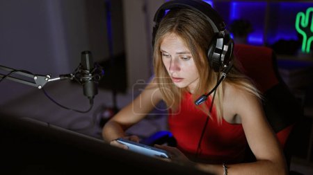 Téléchargez les photos : Focalisé jeune femme portant des écouteurs à l'aide d'une tablette dans une salle de jeux sombre la nuit - en image libre de droit