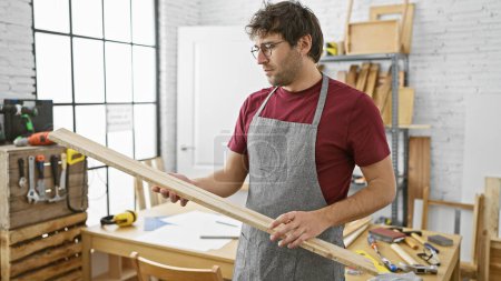Foto de Hombre hispano inspecciona madera en un taller de carpintería soleado con delantal y gafas. - Imagen libre de derechos