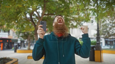 Foto de Emocionado hombre barbudo con teléfono inteligente celebra al aire libre en la ciudad. - Imagen libre de derechos