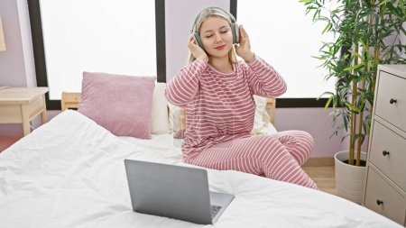Téléchargez les photos : Une jeune femme en pyjama rayé aime la musique sur écouteurs tout en se relaxant dans sa chambre avec un ordinateur portable et une plante d'intérieur. - en image libre de droit