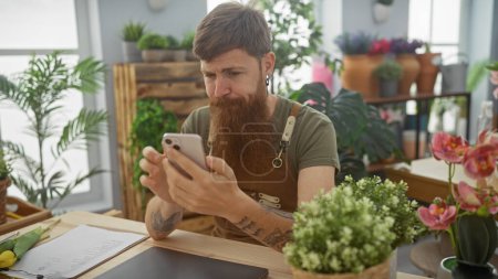 Photo for Bearded man using smartphone in a flower shop surrounded by lush plants - Royalty Free Image