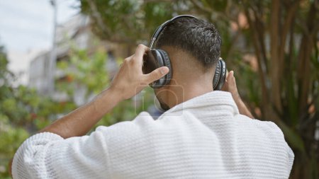 Foto de Un joven disfruta de la música en auriculares al aire libre con una vegetación borrosa en el fondo, ejemplificando el ocio urbano. - Imagen libre de derechos
