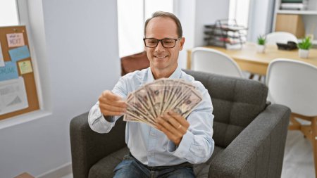 Téléchargez les photos : Homme d'affaires souriant d'âge moyen, comptant avec confiance sa richesse en dollars, assis sur un canapé de bureau dans un cadre intérieur serein - en image libre de droit