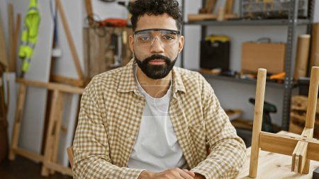 Téléchargez les photos : Beau jeune homme hispanique barbu portant des lunettes de sécurité dans un atelier de menuiserie - en image libre de droit