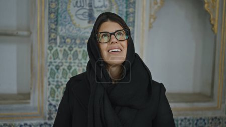 A joyful woman in glasses wearing a hijab stands in an ornate turkish palace, featuring intricate tile work.