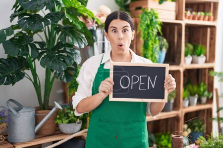 Foto de Young hispanic woman working at florist holding open sign scared and amazed with open mouth for surprise, disbelief face - Imagen libre de derechos