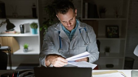 Focused hispanic man in lab coat reviewing documents in a modern clinic office at night.