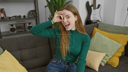 Foto de Una joven alegre hace un gesto aceptable sobre su ojo, sentado alegremente en un ambiente interior acogedor. - Imagen libre de derechos