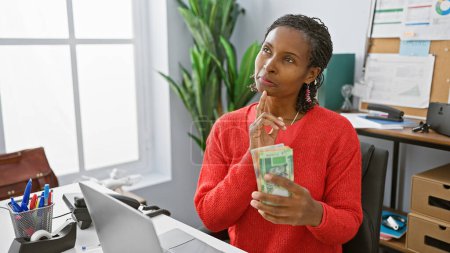 Una mujer reflexiva en un suéter rojo sostiene rands sudafricanos en una oficina, ponderando decisiones financieras.