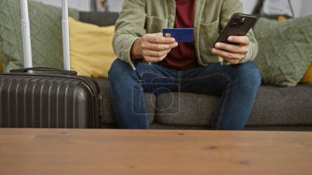 Photo for A young man sitting indoors with a suitcase, holding a credit card and smartphone, possibly preparing for travel or online shopping. - Royalty Free Image