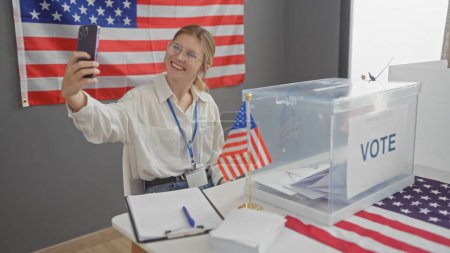 Téléchargez les photos : Une jeune femme souriante prend un selfie avec un smartphone dans un centre de vote américain orné d'un drapeau américain. - en image libre de droit