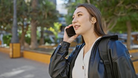 Foto de Una mujer joven habla en un teléfono inteligente mientras camina por una calle urbana soleada y arbolada. - Imagen libre de derechos