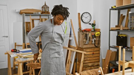 Photo for A young african american woman with dreadlocks wearing an apron stands thoughtfully in a carpentry workshop. - Royalty Free Image