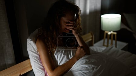 Una mujer caucásica angustiada sentada en la cama con lámpara en el interior por la noche, retratando el estrés o la tristeza.