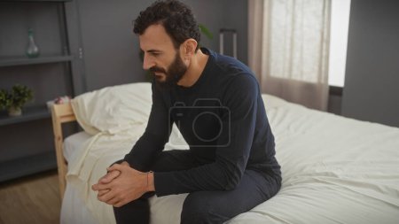 Foto de Un hombre pensativo con barba se sienta solo en una cama en un dormitorio moderno, evocando sentimientos de introspección. - Imagen libre de derechos