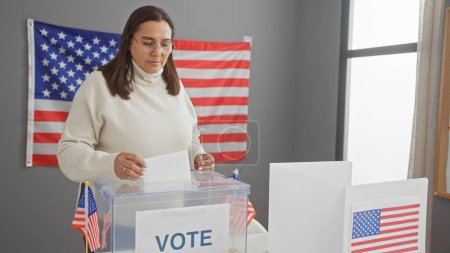 Una mujer hispana de mediana edad emite su voto en un centro electoral de EE.UU. con una bandera americana en el fondo.