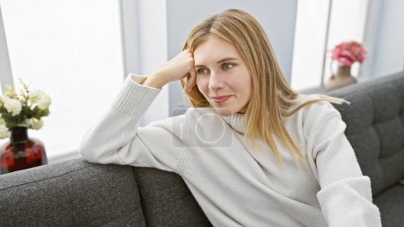 Photo for Blonde woman with blue eyes in a white sweater relaxing on a grey couch indoors, looking thoughtfully aside. - Royalty Free Image