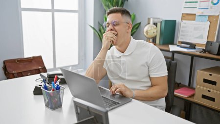 Foto de Un joven hispano con atuendo casual bosteza mientras trabaja con un portátil en una oficina luminosa. - Imagen libre de derechos