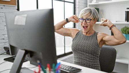 A joyful middle-aged woman with grey hair celebrates success at her office workstation indoors.