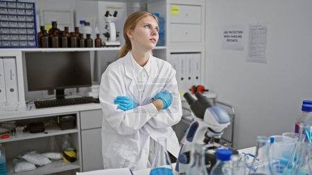 Foto de Mujer joven pensativa con bata de laboratorio con los brazos cruzados de pie en un laboratorio de ciencias en el interior. - Imagen libre de derechos