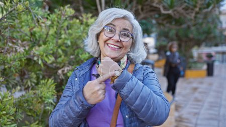 Téléchargez les photos : Une femme mûre souriante avec des lunettes et des pointes de cheveux gris à sa montre dans un cadre vert parc extérieur. - en image libre de droit