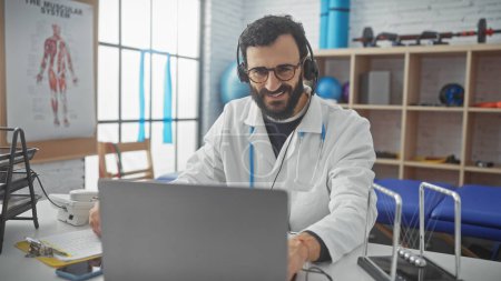 Photo for Smiling bearded man wearing labcoat and headphones working on laptop in physical therapy clinic - Royalty Free Image