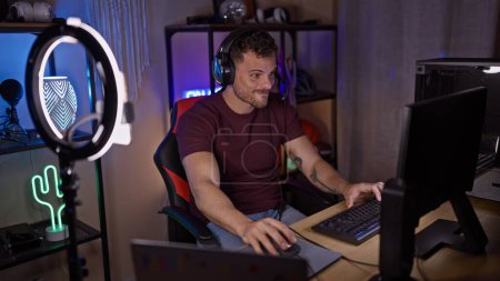 Téléchargez les photos : Un jeune homme hispanique concentré avec une barbe utilise un ordinateur dans une salle de jeux faiblement éclairée la nuit. - en image libre de droit