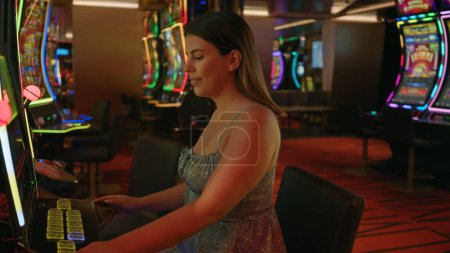 Téléchargez les photos : Une jeune femme brune hispanique adulte s'engage avec une machine à sous dans un cadre de casino, exsudant un air de succès plein d'espoir. - en image libre de droit