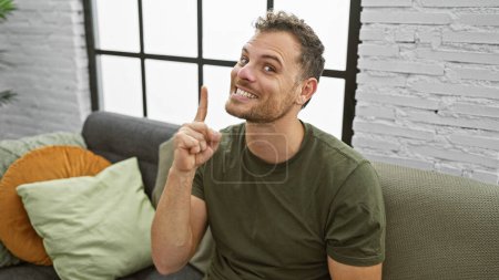 Foto de Un joven alegre hispano con barba, con una camisa casual, haciendo un gesto de una idea mientras está sentado en un sofá en el interior. - Imagen libre de derechos