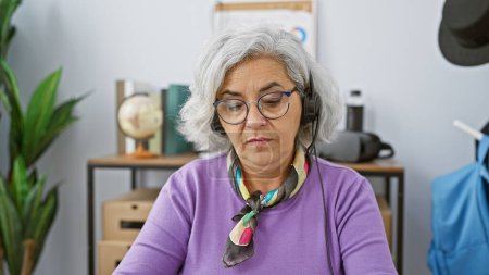 Photo for Portrait of a focused, grey-haired woman wearing headphones in a modern office setting. - Royalty Free Image