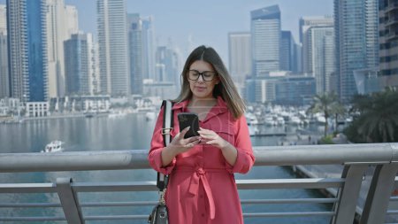 Brünette Frau in Rot nutzt Smartphone vor Skyline von Dubais Yachthafen und verkörpert Luxus, Reisen und Modernität.