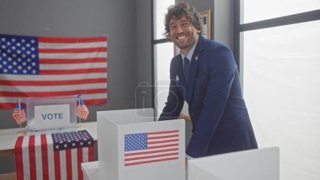Un joven hispano sonriente con una barba vestida con un traje está junto a una cabina de votación con un telón de fondo de bandera americana en un entorno interior.