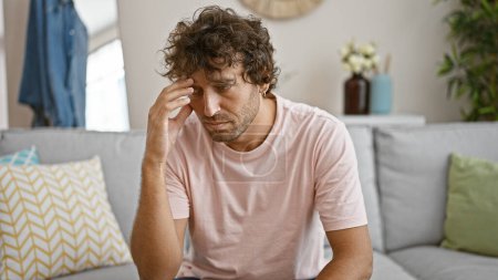 Foto de Hombre hispano joven preocupado sentado en casa en un sofá, mostrando síntomas de estrés, contemplación o dolor de cabeza. - Imagen libre de derechos
