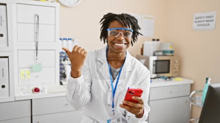 Foto de Mujer afroamericana joven con rastas y bata de laboratorio en un laboratorio, sonriendo mientras sostiene un teléfono inteligente. - Imagen libre de derechos