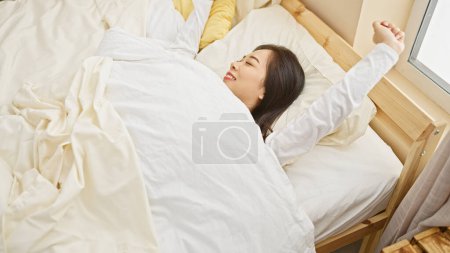 Photo for A young asian woman stretching with joy in a sunlit bedroom after waking up. - Royalty Free Image