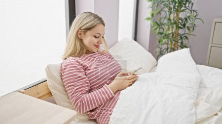 Une jeune femme blonde en pyjama rayé se détend dans une chambre avec un smartphone.