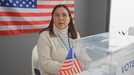 Eine reife hispanische Frau beaufsichtigt die Stimmabgabe in einem US-Wahlzentrum, im Hintergrund eine amerikanische Flagge.