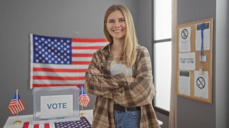 Blonde Frau lächelt selbstbewusst mit verschränkten Armen in einem US-Wahlzentrum mit Wahlurne und amerikanischer Flagge.