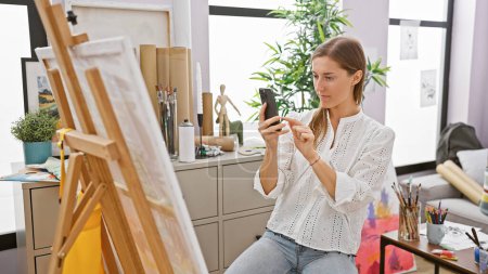 Foto de Una mujer caucásica enfocada artista en un estudio utiliza un teléfono inteligente al lado de su caballete y suministros de arte. - Imagen libre de derechos