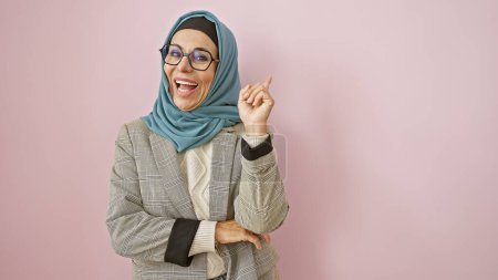 Téléchargez les photos : Une femme joyeuse d'âge moyen en hijab et lunettes pose sur un fond rose, affichant confiance et bonheur. - en image libre de droit